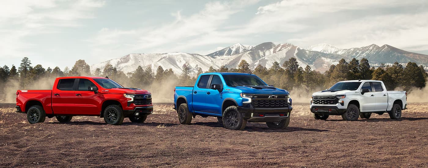 A red, a blue, and a white 2024 Chevy Silverado 1500 are parked in an open dirt lot.