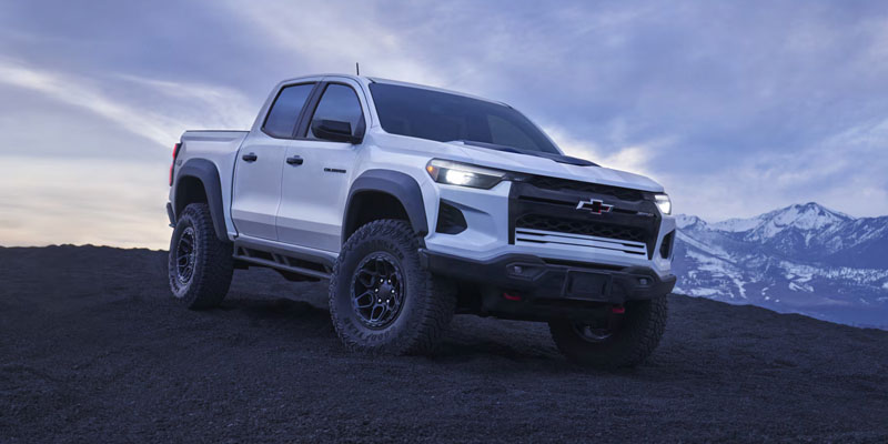White 2026 Chevy Colorado pickup truck parked on rugged terrain in Lafayette, LA, featuring off-road tires, a bold front grille, and a mountainous landscape in the background under a cloudy sky.